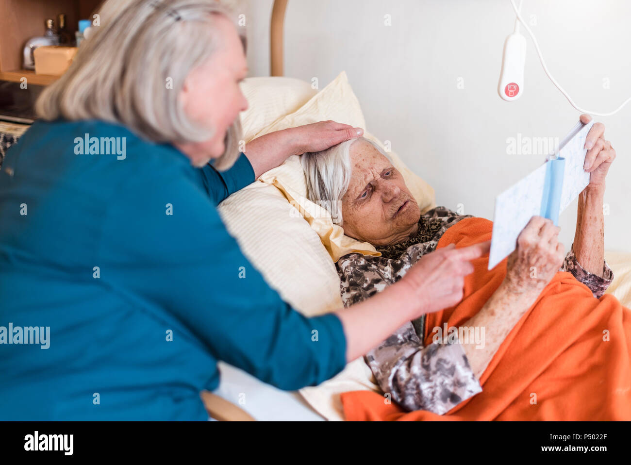 Woman taking care of old woman lying in bed reading book Stock Photo