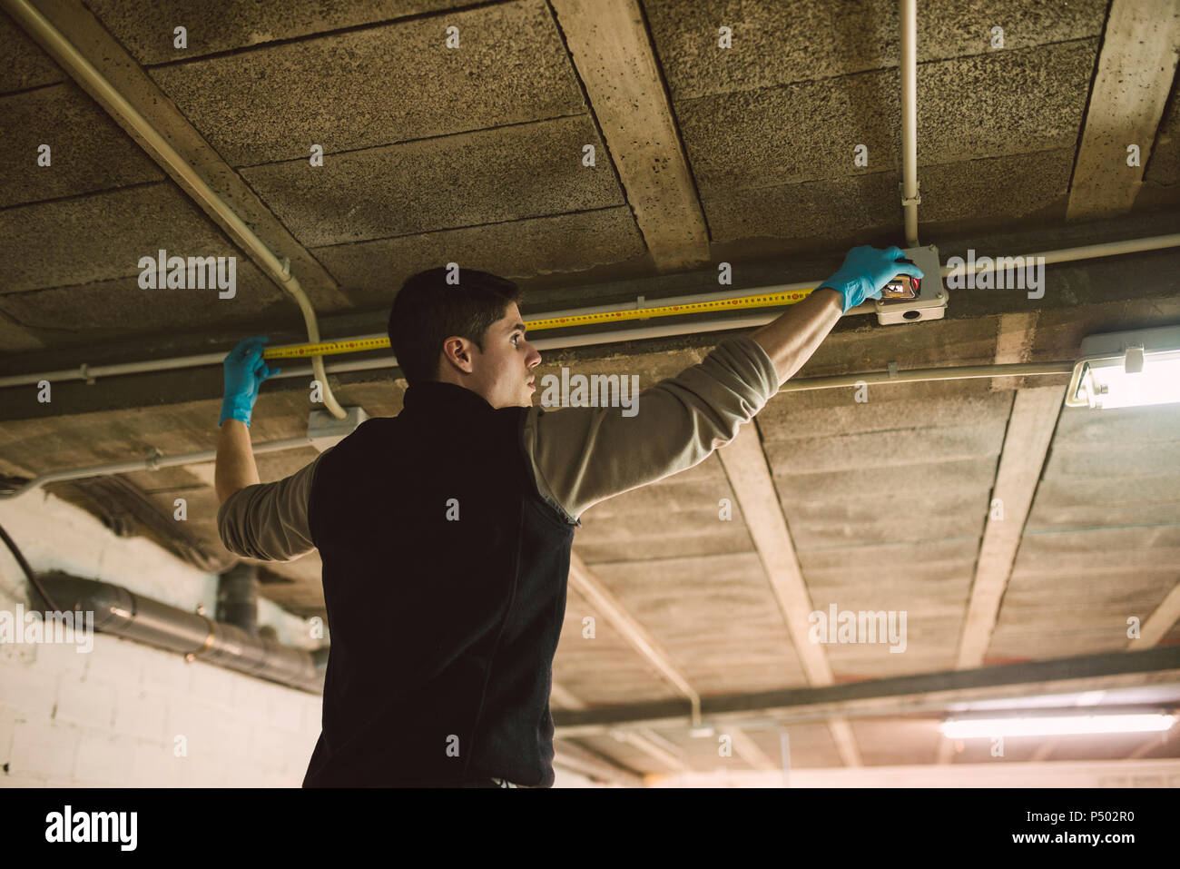 Man working on conduit at the ceiling measuring with a tape measure Stock Photo