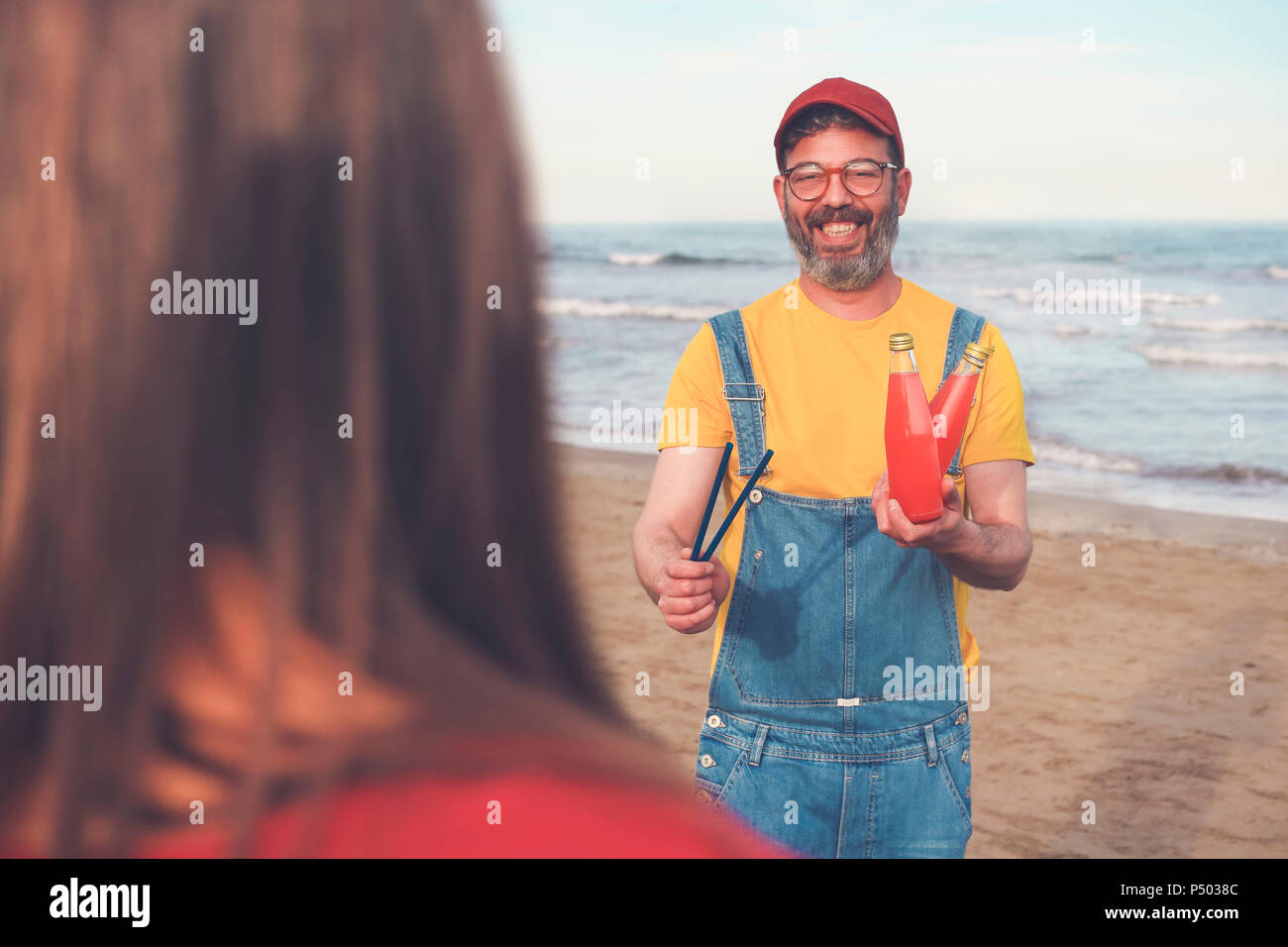 Happy man in dungarees on the beach offering soft drink to woman Stock Photo