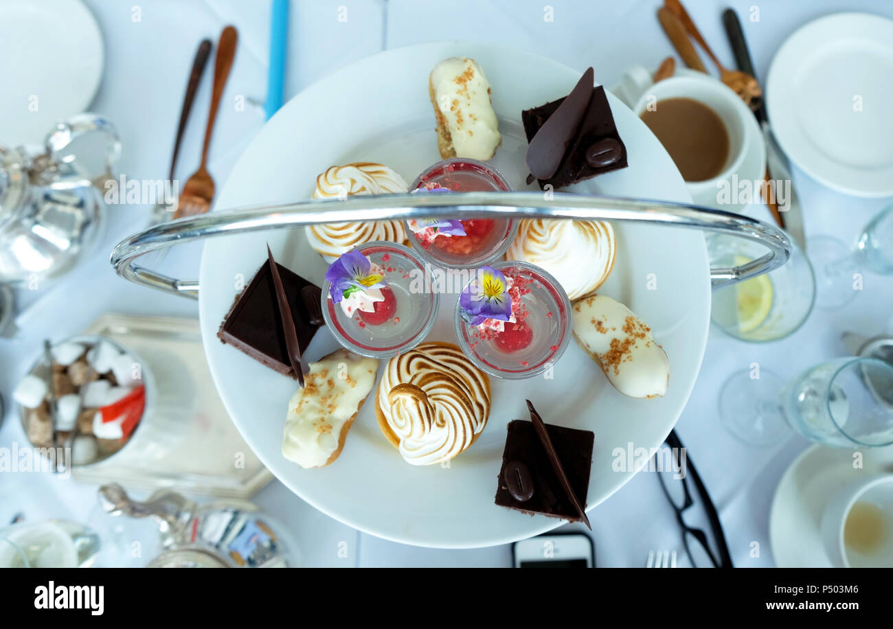 looking down onto a table with a English afternoon tea on it, a round plate of colourful cakes with silver tea and coffee pots and white cups white li Stock Photo