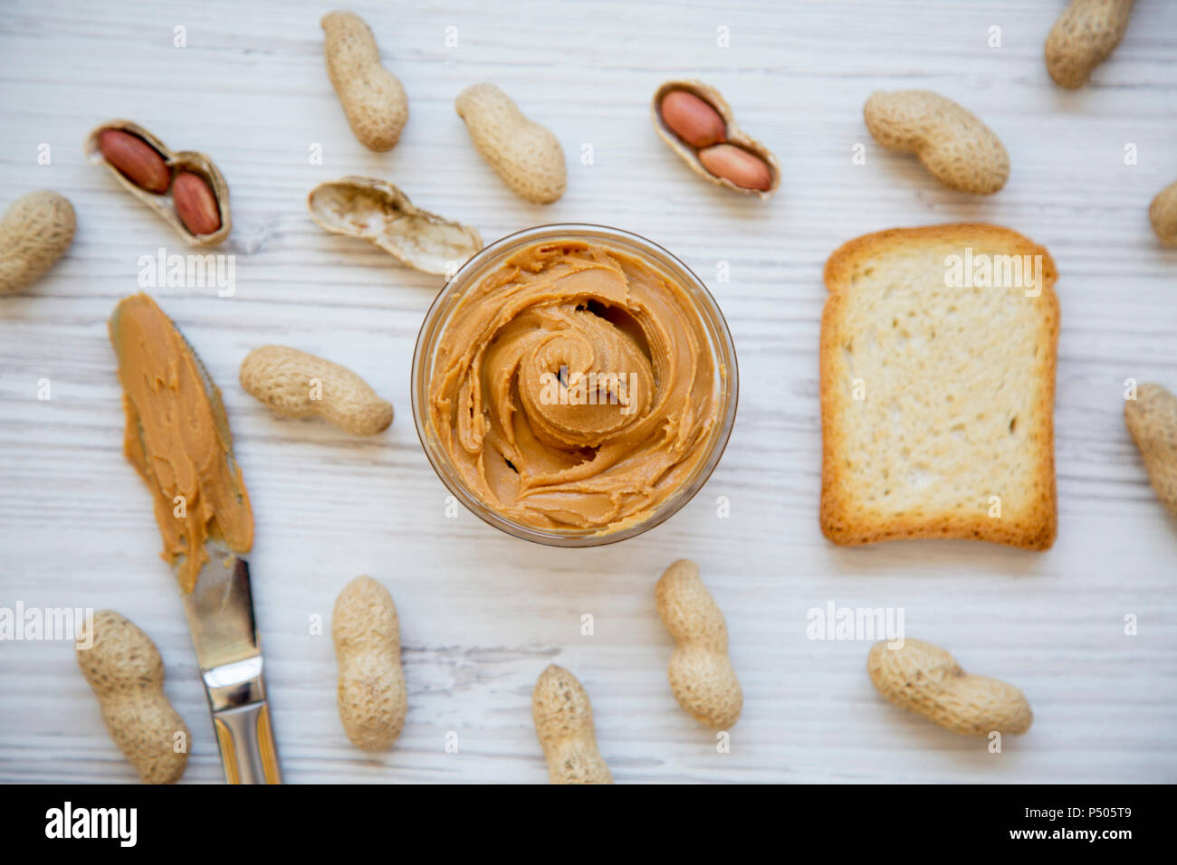 Toast, bowl of peanut butter, knife and peanuts in shells on a white wooden background, top view. From above. Stock Photo