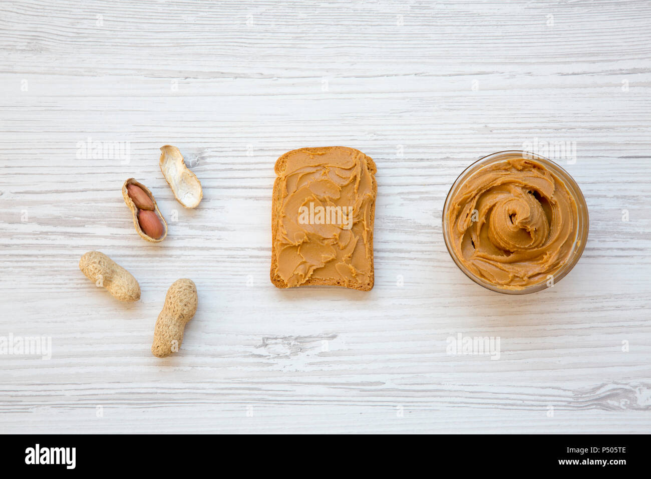 Toast with peanut butter, bowl of peanut butter and peanuts in shells on a white wooden background, top view. Flat lay. Stock Photo