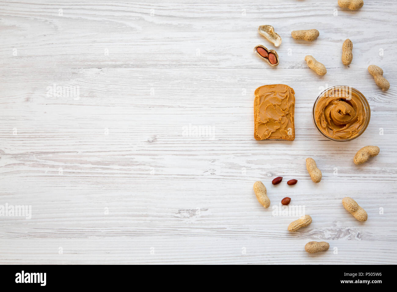 Toast, bowl of peanut butter and peanuts in shells on a white wooden background, top view. Copy space. From above. Stock Photo