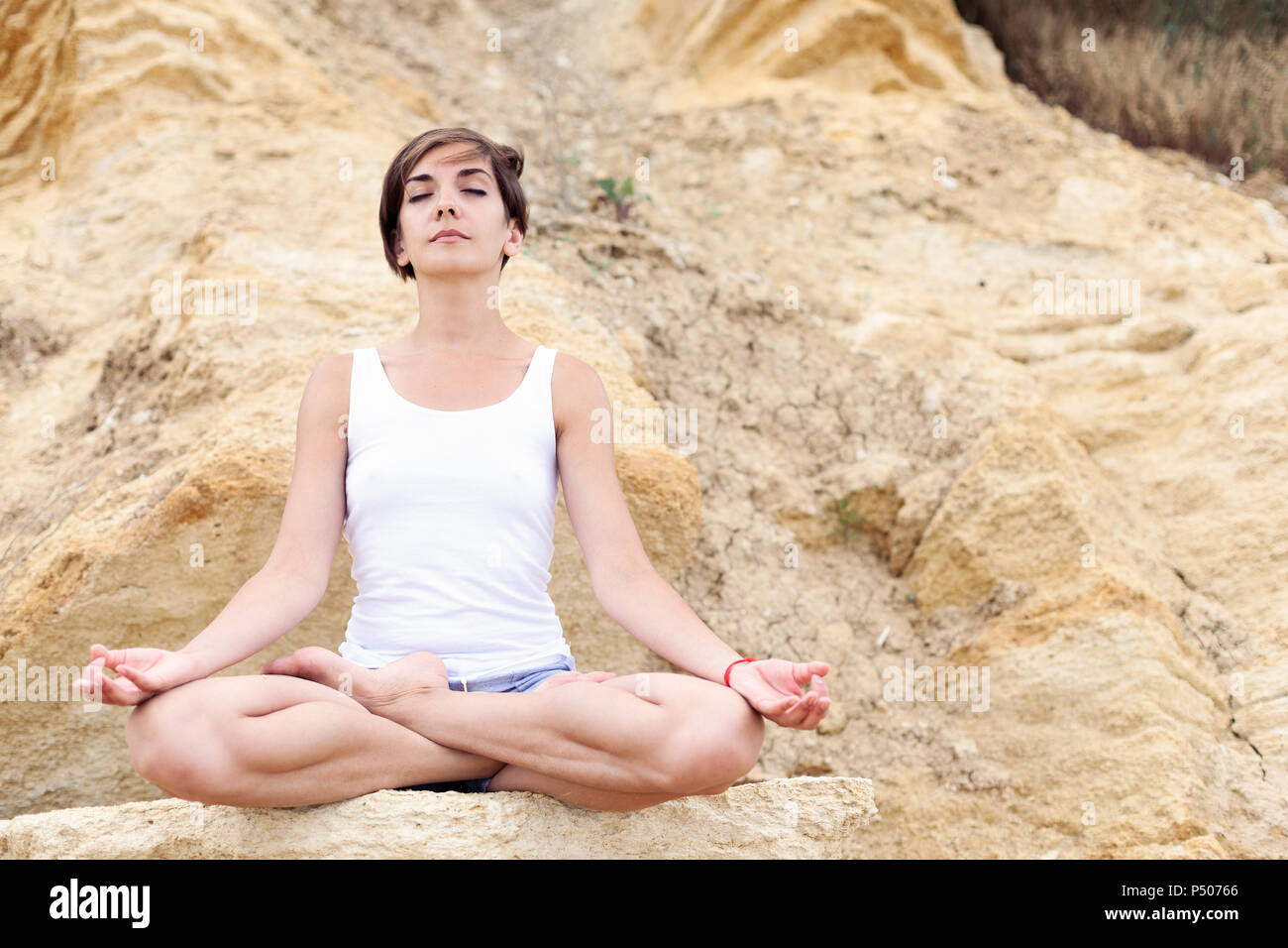 A beautiful young girl with short hair is dressed in shorts and a white  jersey is practicing yoga on the background of rocks. Pose of the lotus.  The c Stock Photo 