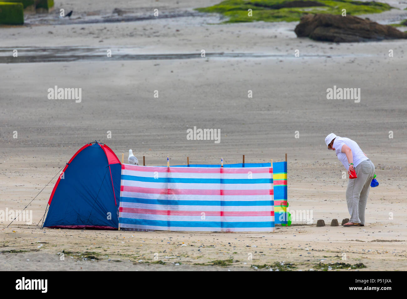 An elderly woman wearing a sunhat making sand castles in the sand alone at looe beach in Cornwall with a tent and wind breaker Stock Photo