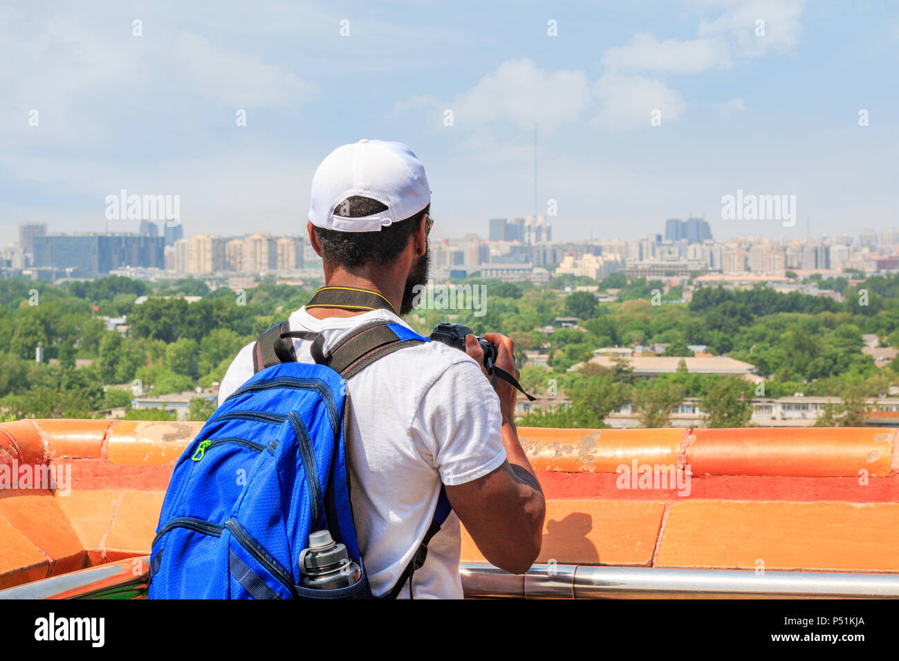 A male traveler in a white T-shirt and cap makes a photo of the city landscape from the gazebo of Jingshan Park. Stock Photo