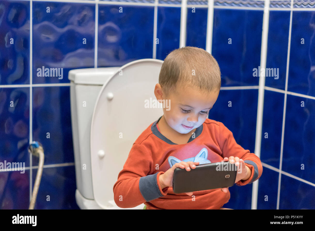Little boy sitting on the toilet in the bathroom at home with using a smartphone. Toddler boy sitting on toilet with smartphone. Stock Photo