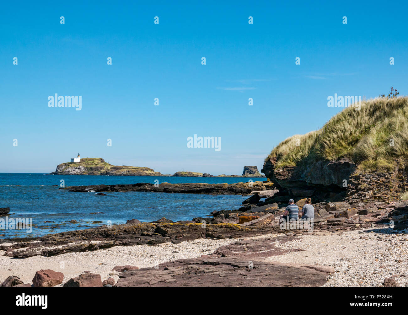 John Muir Way, East Lothian, Scotland, UK, 24th June 2018. UK Weather: A warm sunny day during the 2018 Summer heatwave brought people to the coast for leisure pursuits. A couple take a rest on the rocky shore with a view of the lighthouse on Fidra Island, reputedly the inspiration for Robert Louis Stevenson’s novel Treasure Island Stock Photo