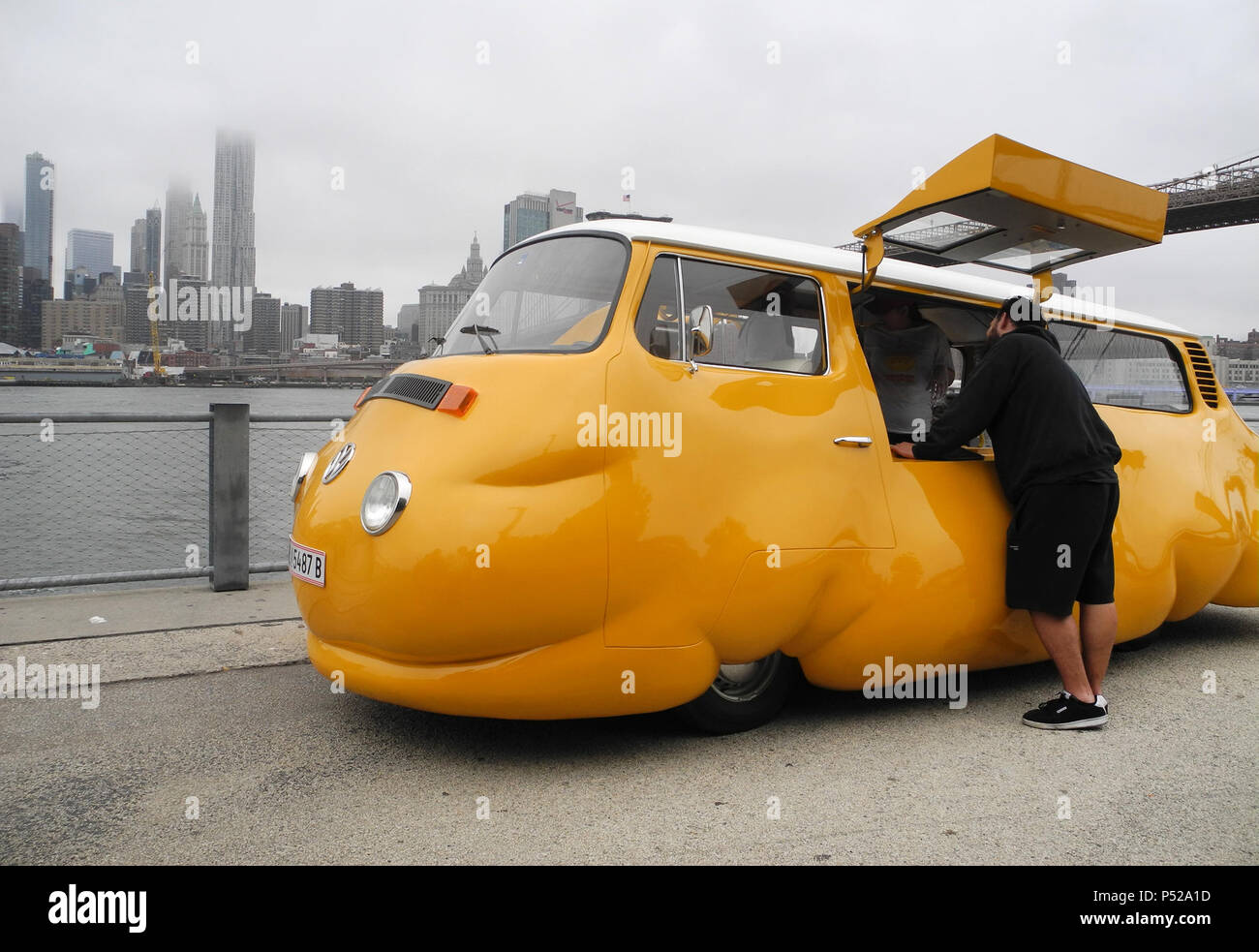 New York, USA. 23rd June, 2018. From a mustard-yellow bus, sausages are distributed free of charge - in the background of the 'Hot Dog Bus' by the Austrian artist Erwin Wurm, the skyline of Brooklyn can be seen across the river. The baggy, chubby minibus in the Brooklyn neighborhood is reminiscent of Wurm's Fat Car series of fat cars. Credit: Johannes Schmitt-Tegge/dpa/Alamy Live News Stock Photo
