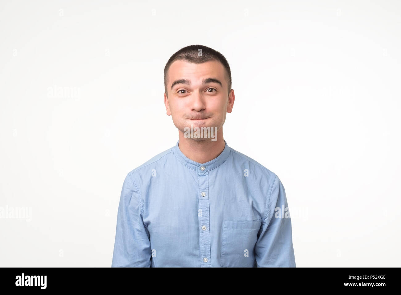 Funny young man in blue shirt grimacing, staring at camera nflating cheeks, holding his breath, trying hard not to laugh. Try to hold emotion inside Stock Photo
