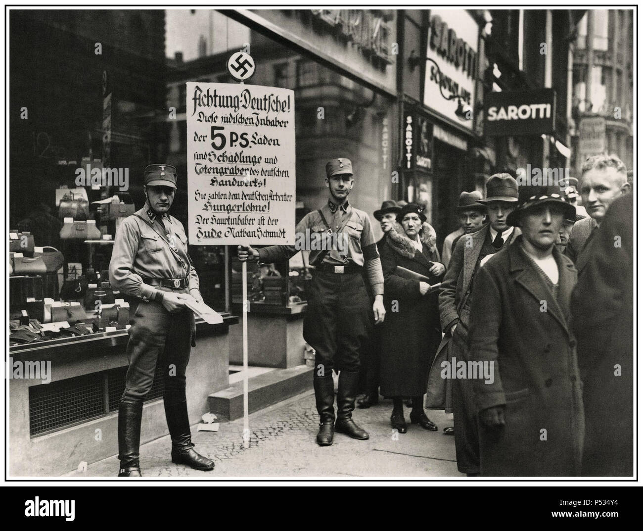 Vintage 1930’s anti-Jewish racist inflammatory image of SA Sturmbleitung or “Brownshirts” storm detachment members calling for the boycott of Jewish shops in Friedrichstraße, Berlin; April 1, 1933. The sign with Nazi swastika emblem says: “Germans, Attention! This shop is owned by Jews. Jews damage the German economy and pay their German employees starvation wages. The main owner is the Jew Nathan  Schmidt.” Somber people walk past the shop front. Stock Photo