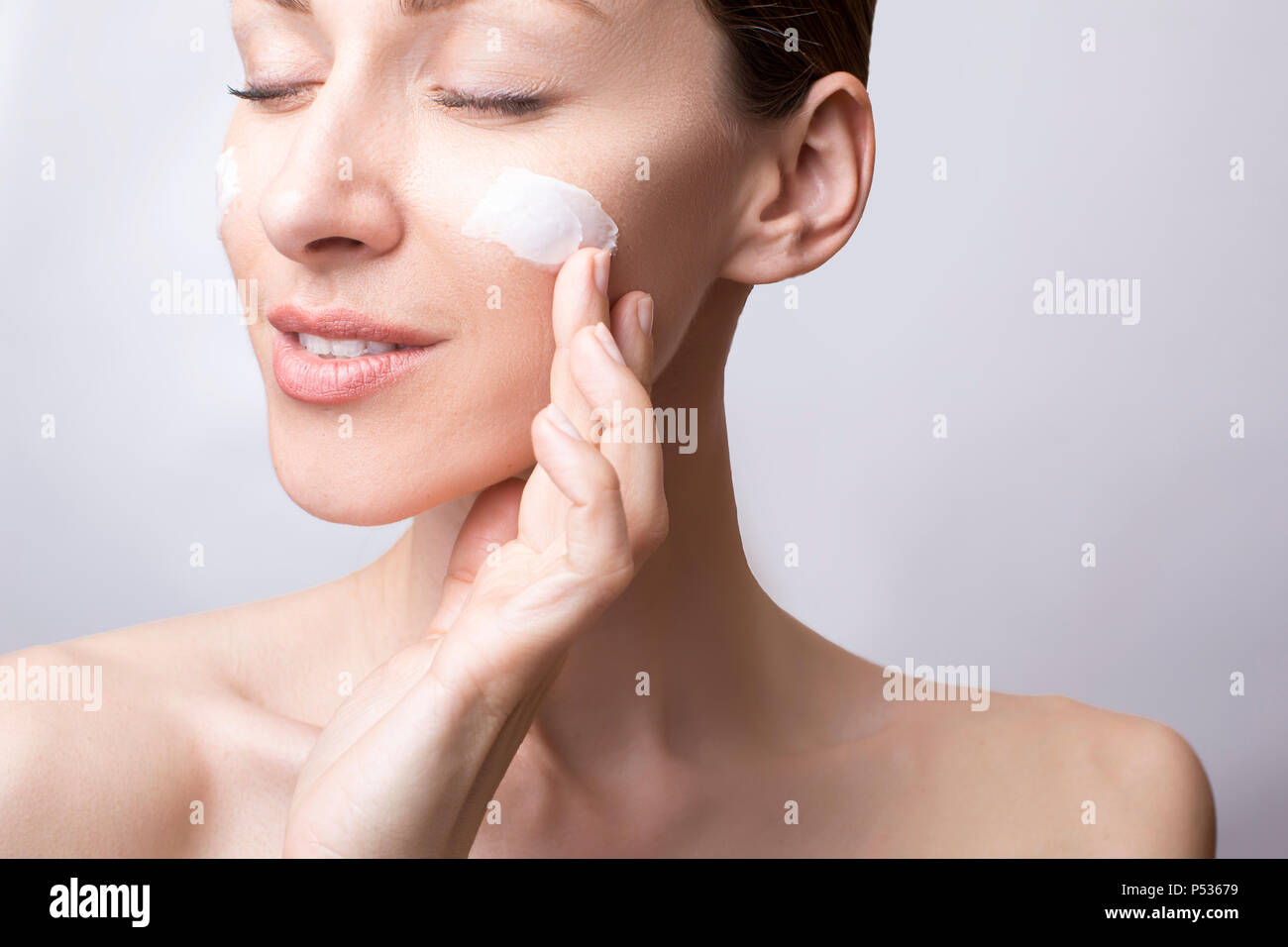 Laughing girl applying moisturizing cream on her face. Photo of young girl with flawless skin on grey background. Skin care and beauty concept Stock Photo