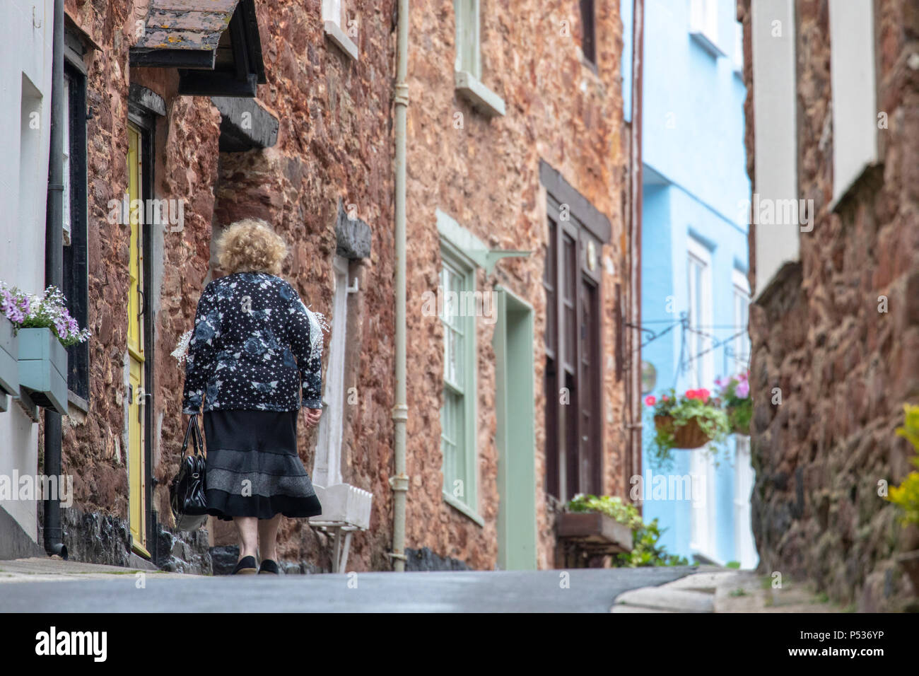 Elderly retired lady walking uphill along a narrow fishing village lane at at the coastal village of Kingsand, Cornwall Stock Photo