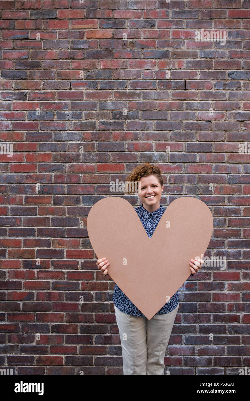 Smiling young woman holding a large heart while standing outside Stock Photo