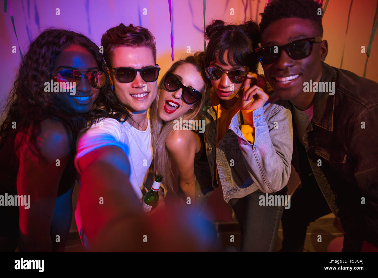 Friends posing for a selfie wearing dark sunglasses at a house party. Young men and women having fun at a colorful house party. Stock Photo