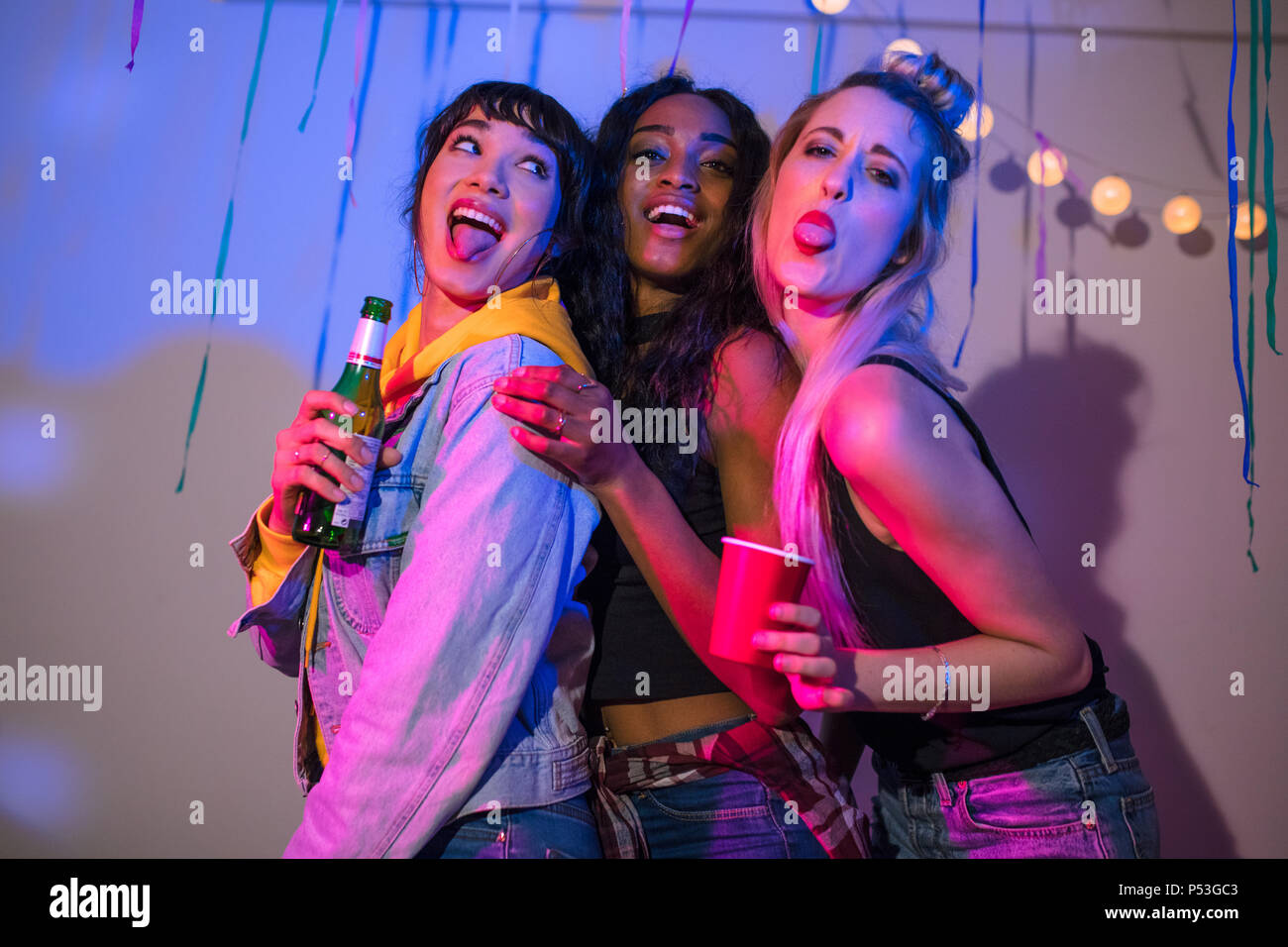 Three young women standing together making faces while holding drinks at a house party. Friends posing for a photograph sticking out their tongues and Stock Photo