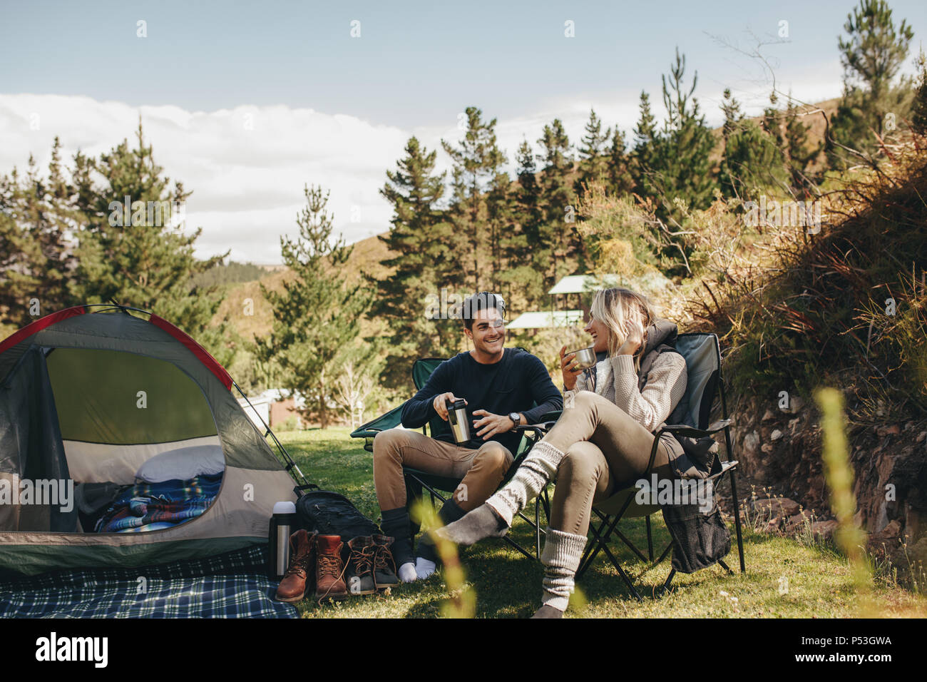 Man and woman sitting in chairs outside the tent having coffee and talking. Couple camping in forest. Stock Photo