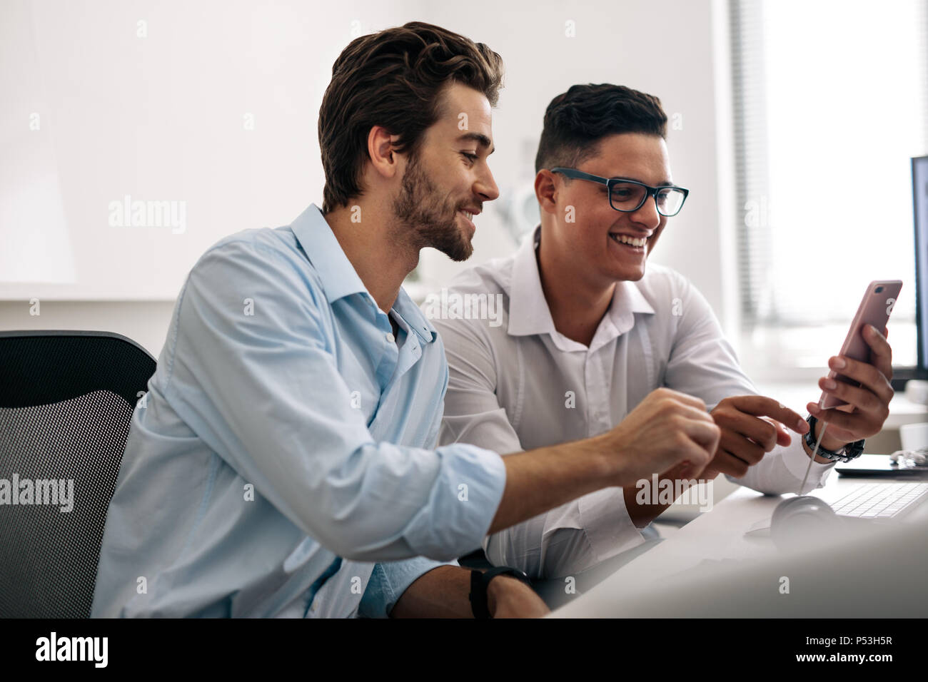 Application developers testing application on mobile phone in  office. Smiling office colleagues sitting at office desk looking at a mobile phone. Stock Photo