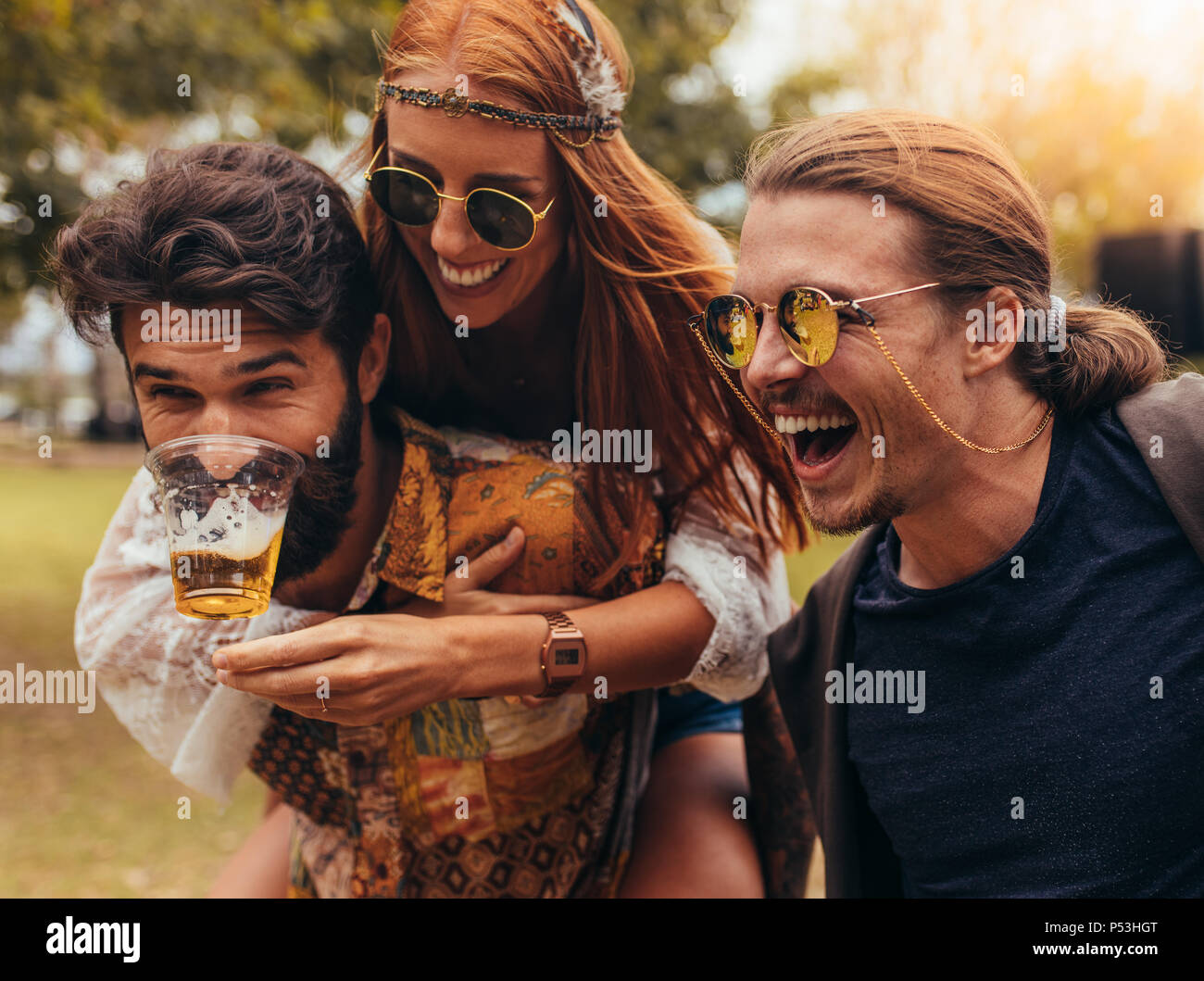 Cheerful young people having fun outdoors at park. Men and woman piggybacking and having beer. Stock Photo