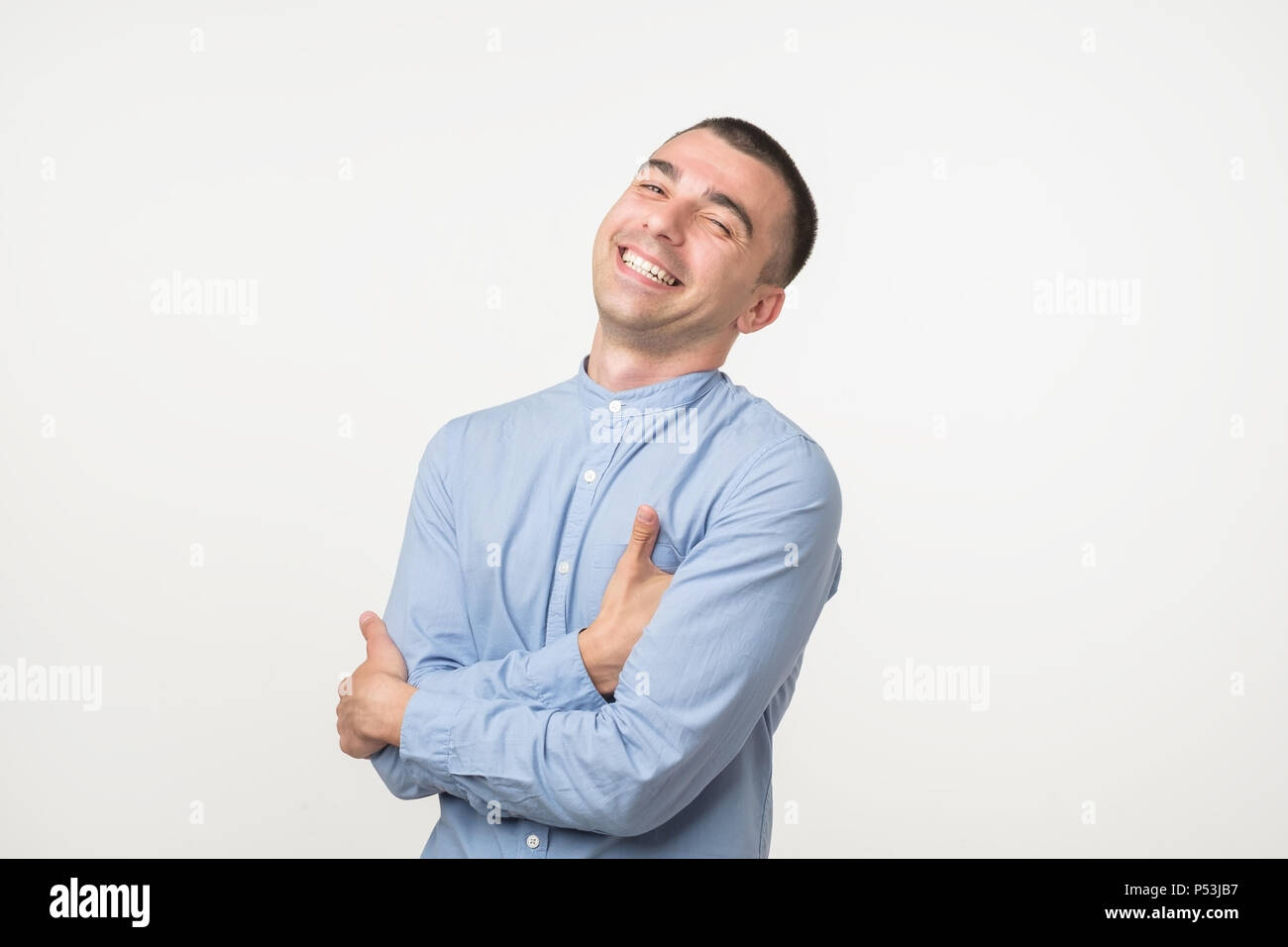 Attractive man in blue shirt laughs at funny story from friend with crossed hands and closed eye Stock Photo