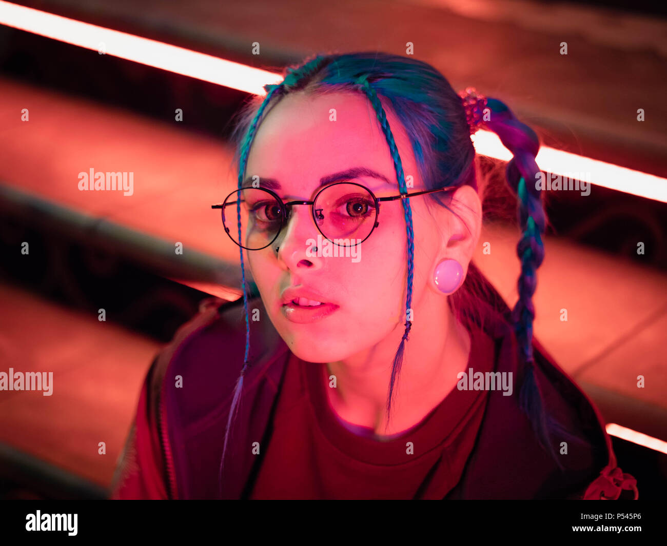 Young pretty girl with unusual hairstyle sitting on stairs which glowing red neon lights. City at night. Dyed blue hair in braids. Happy hipster teenager in glasses. Stock Photo