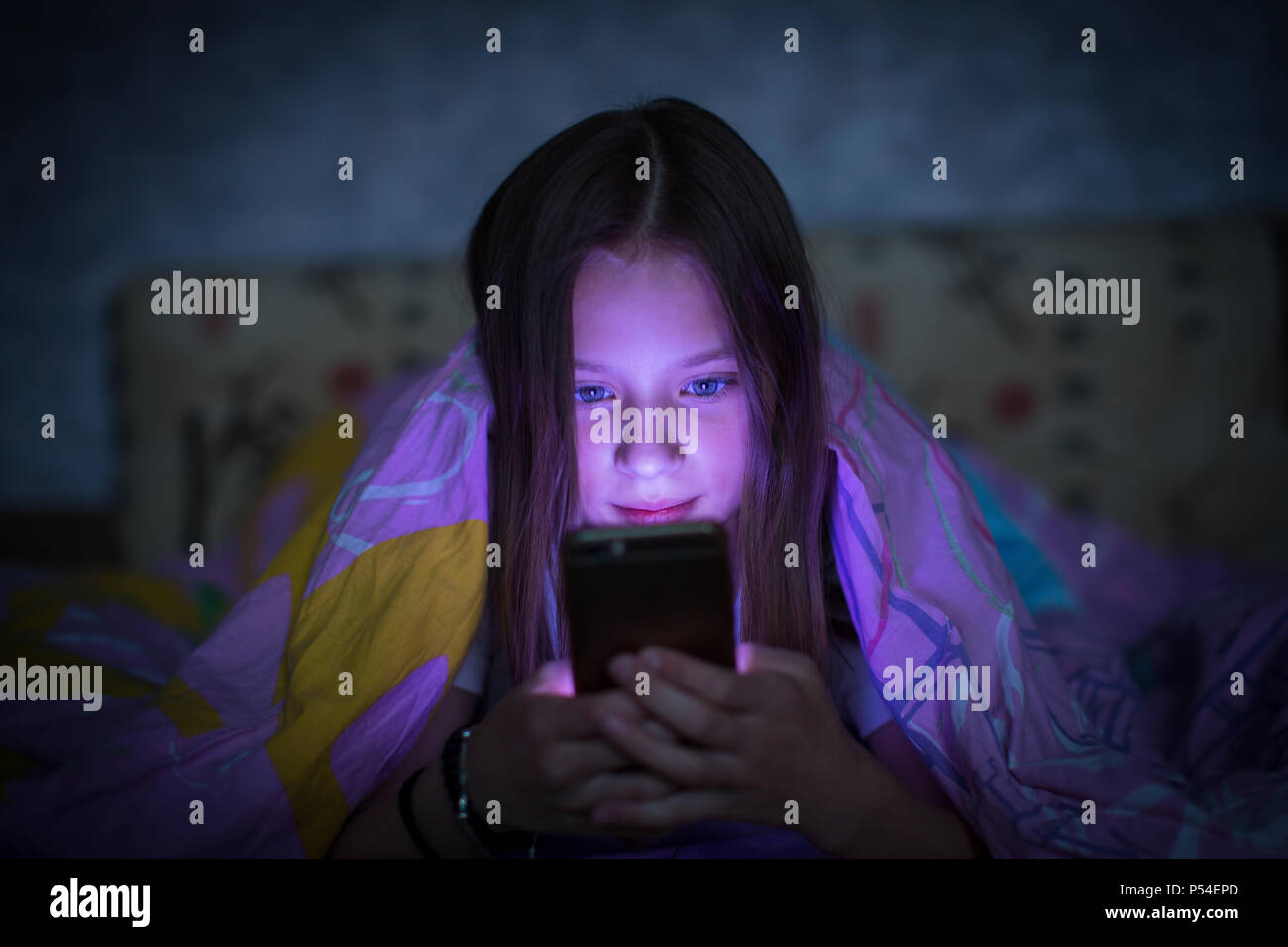 Teen girl lying in bed at night and looking at the glowing screen of smartphone. Stock Photo