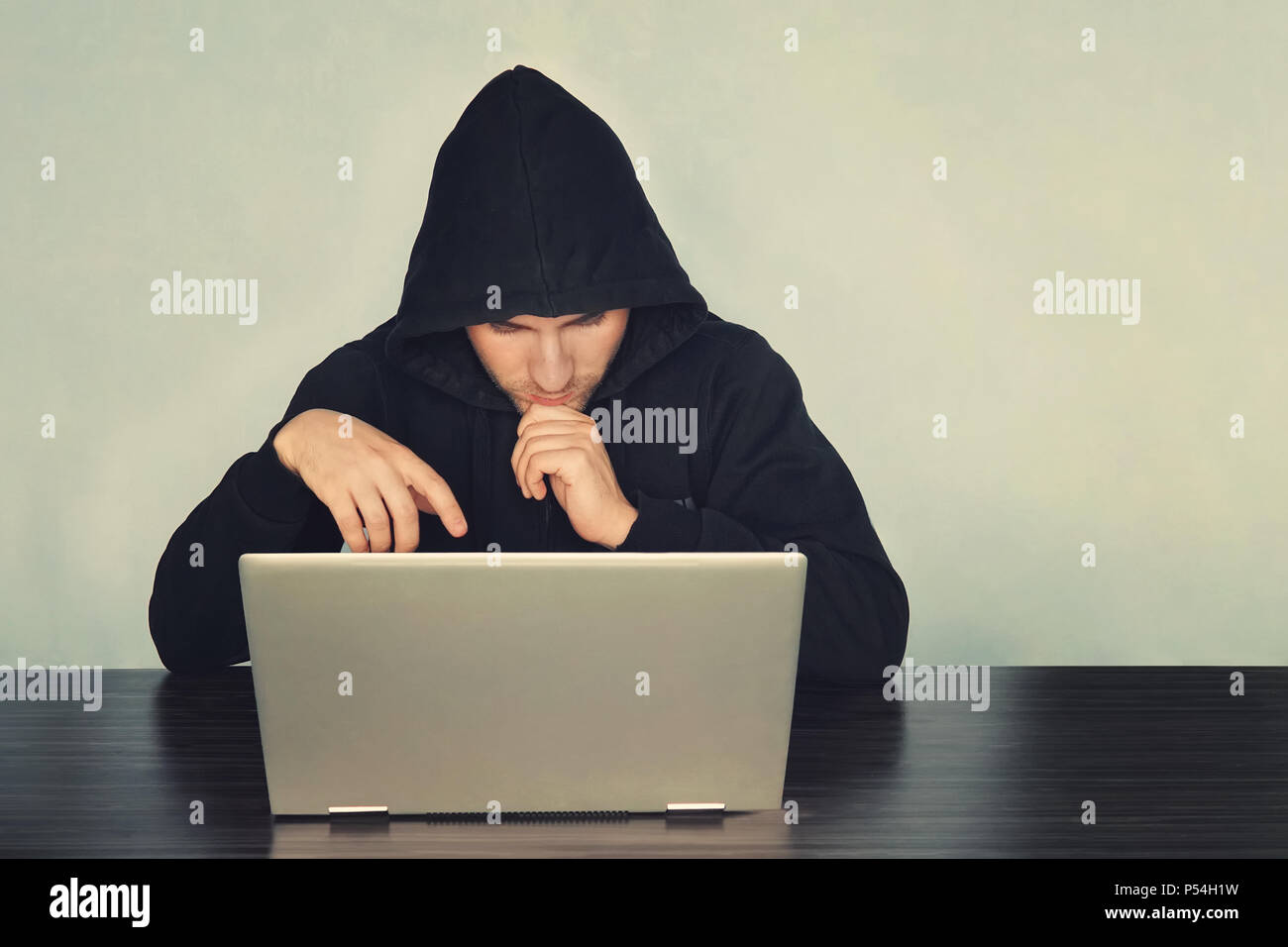 Black masked hacker in the black long sleeve shirt is sitting on the desk in front of laptop and thinking somethings. He is looking at laptop. Blur ba Stock Photo