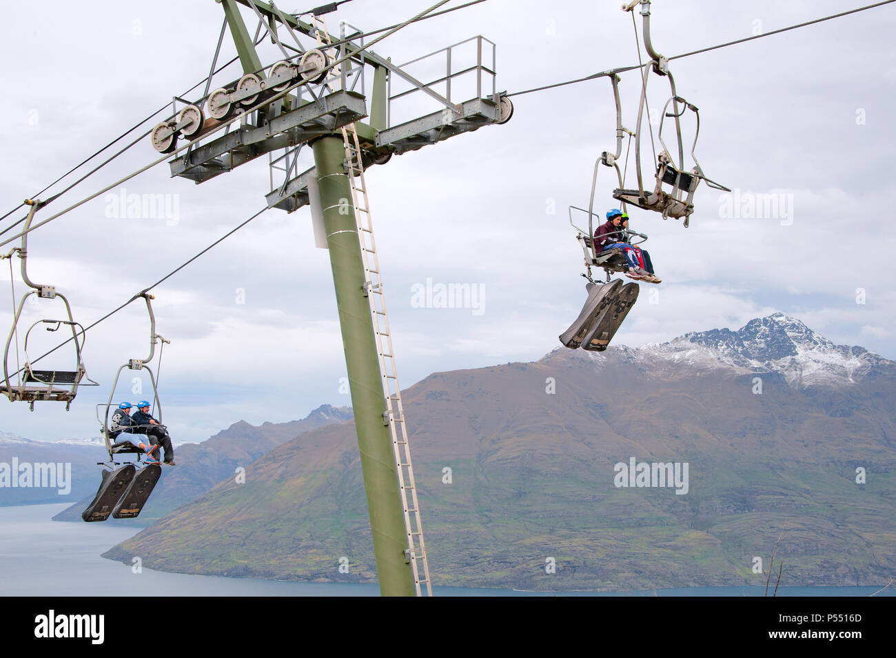 Queenstown, NZ – April 15, 2018: Fun riders with their carts on a cable chair lift going up the hill top for a luge ride with views of Lake and hills. Stock Photo