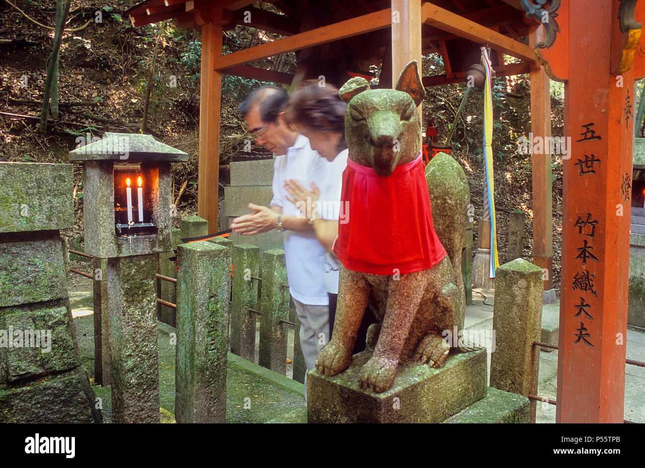 Couple praying, and Kitsune fox statue, at Fushimi Inari-Taisha sanctuary,Kyoto, Japan Stock Photo