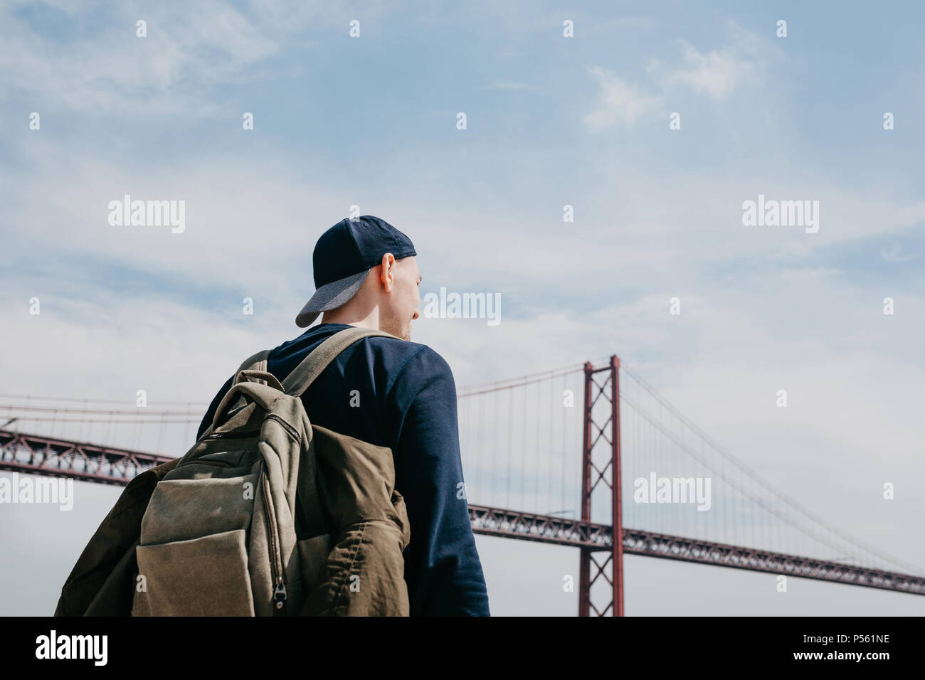 A young traveler or a tourist with a backpack on the waterfront in Lisbon in Portugal next to the 25th of April Bridge Stock Photo