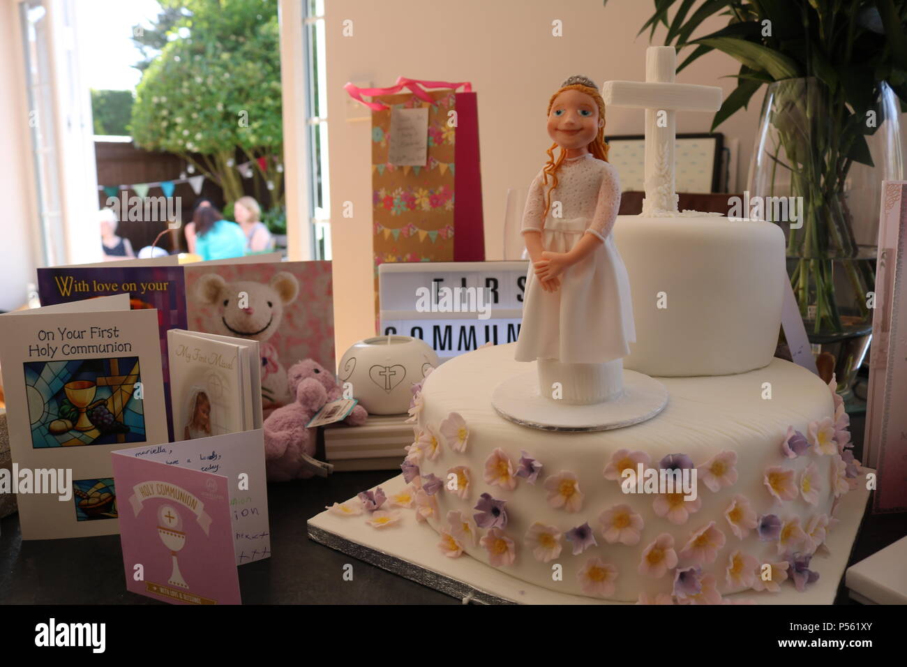 Two tier catholic first communion cake with icing figure of girl and cross, surrounded by cards and gifts with people in background sitting in a garde Stock Photo