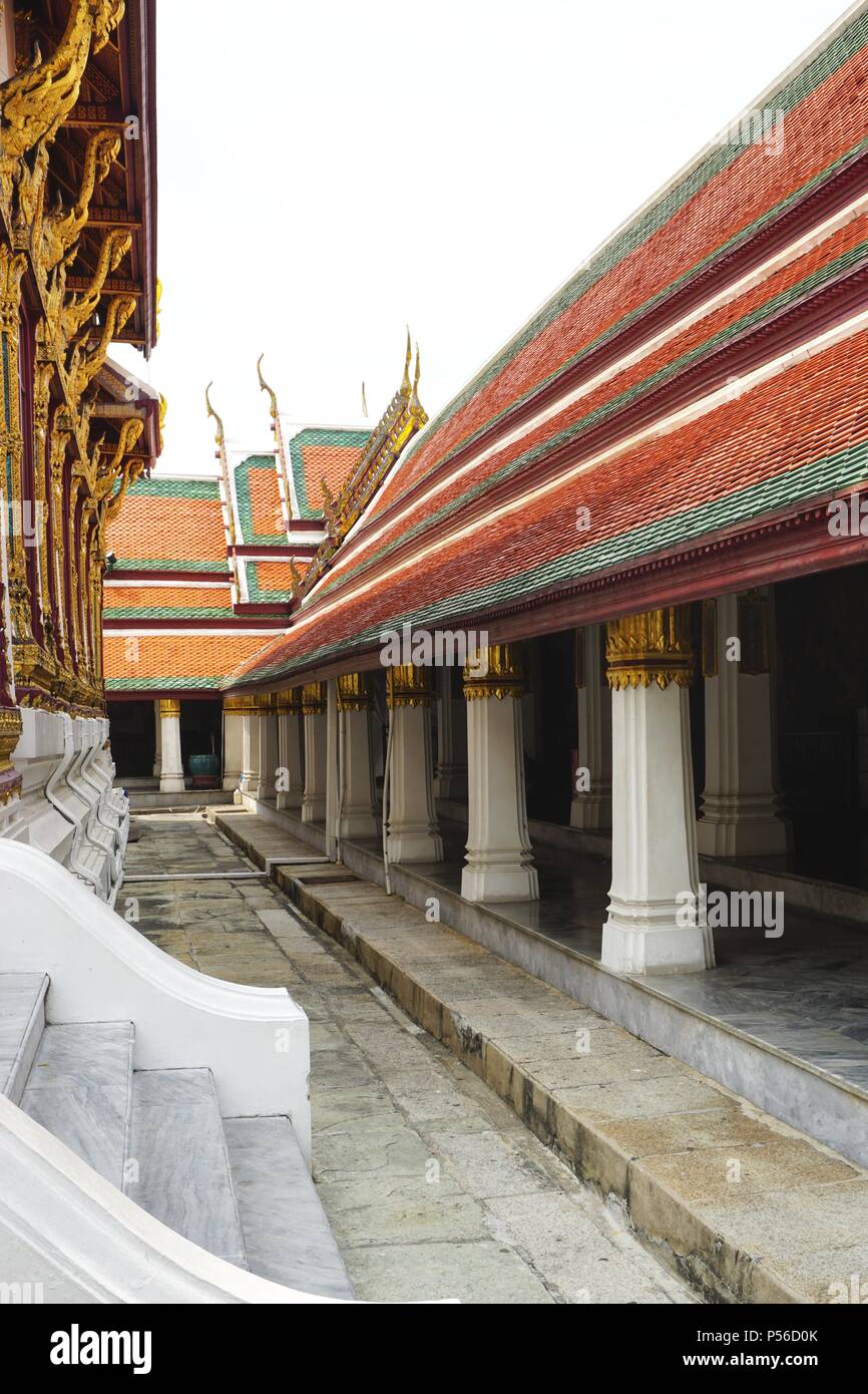 View along a stone walkway at Bangkok imperial palace with matching pillars and colorful tile roof to one side and ornate gilded pillars to the other Stock Photo