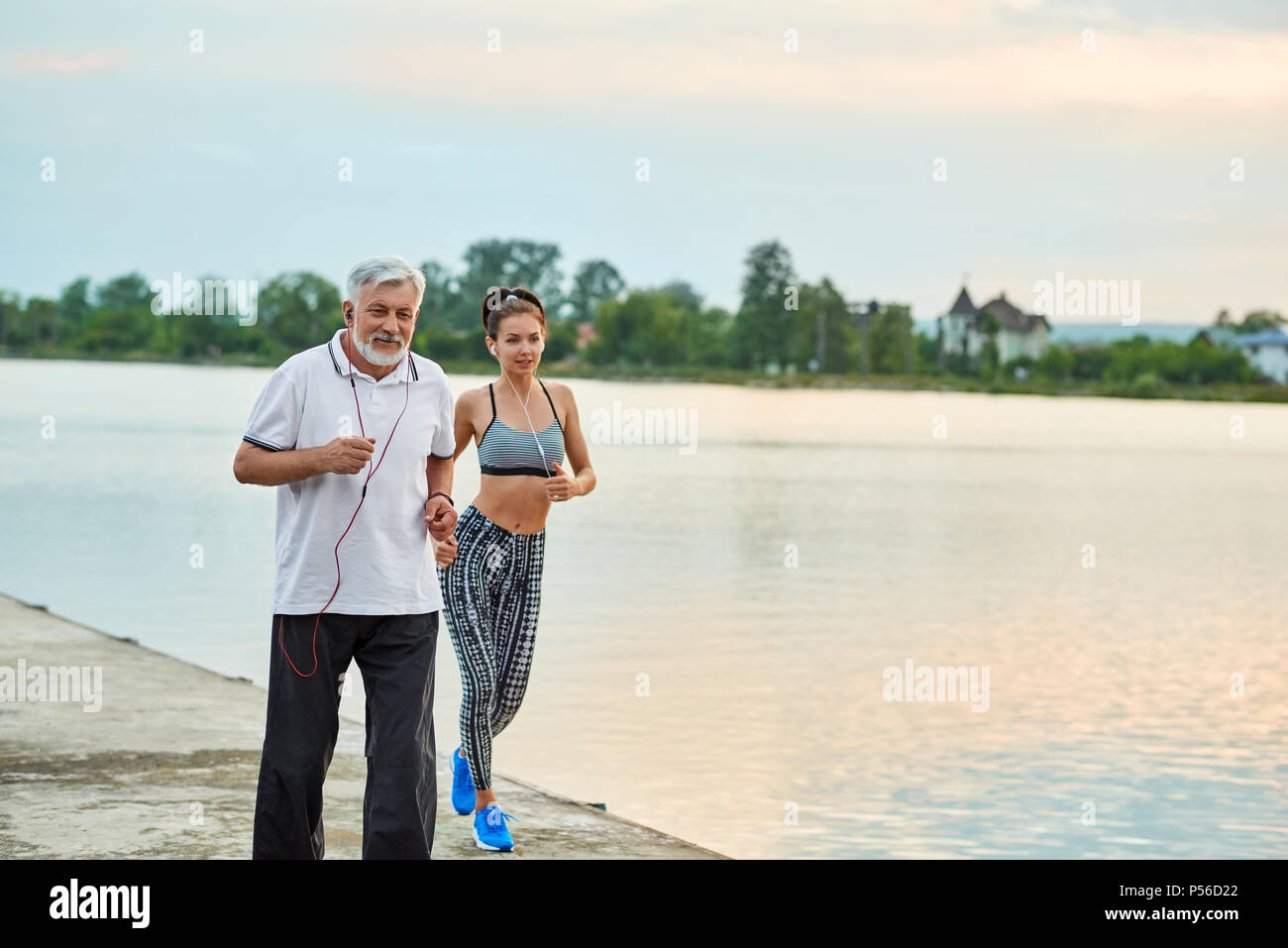 Senior man and active young girl running near city lake. Active lifestyle, healthy body. Sport, yoga, fitness. Different generations. Models having strong, fit figures. Modern sportswear. Stock Photo
