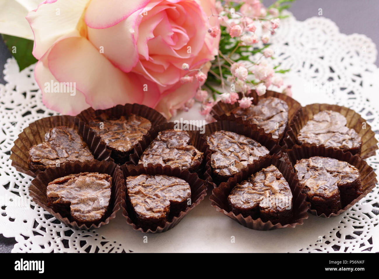 Heart shaped brownies in glassine brown paper cups on a white doily decorated with a white rose with pink borders. Stock Photo
