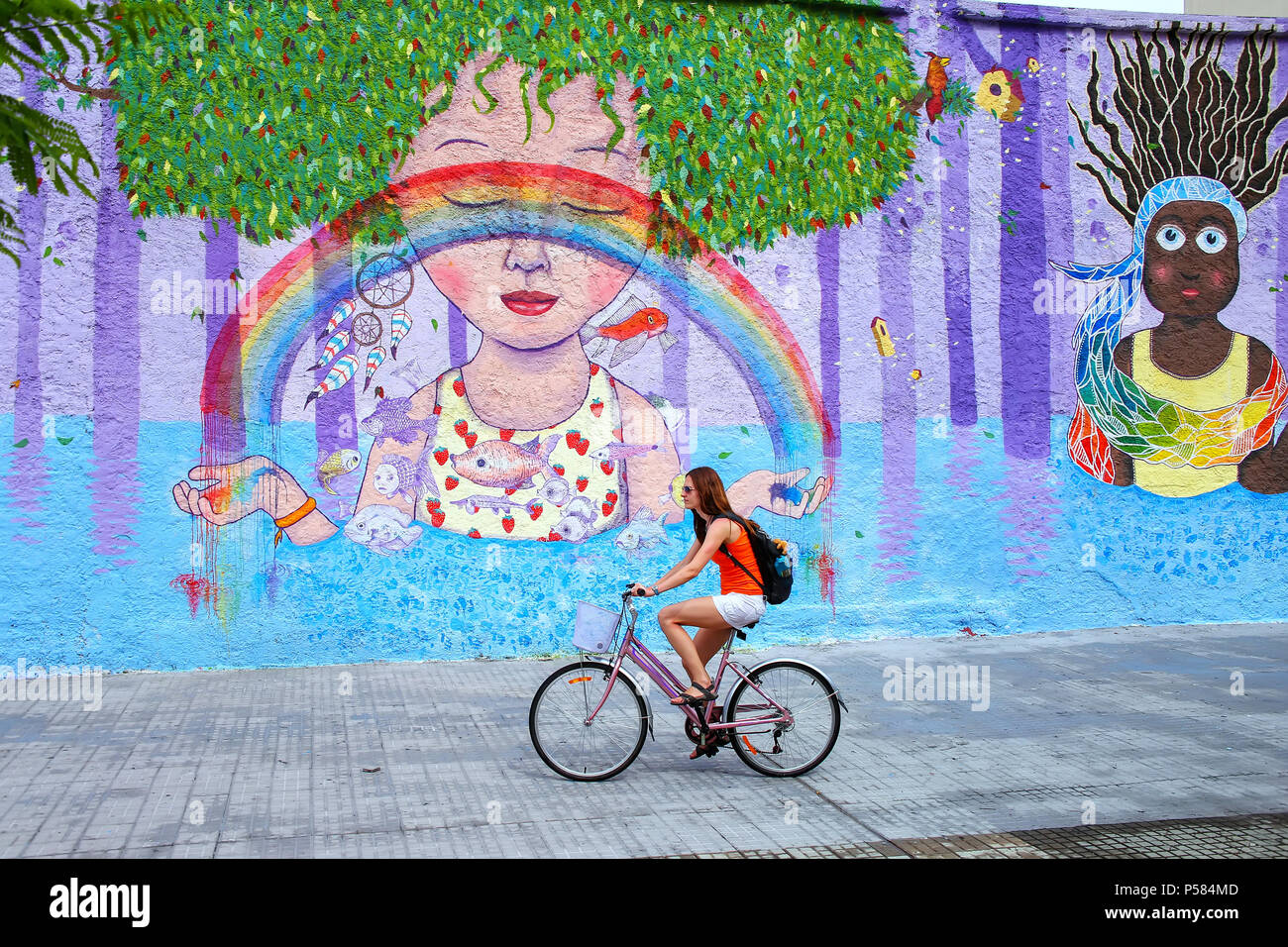Young woman biking along colorful wall in Montevideo, Uruguay. Montevideo is the capital and largest city of Uruguay. Stock Photo