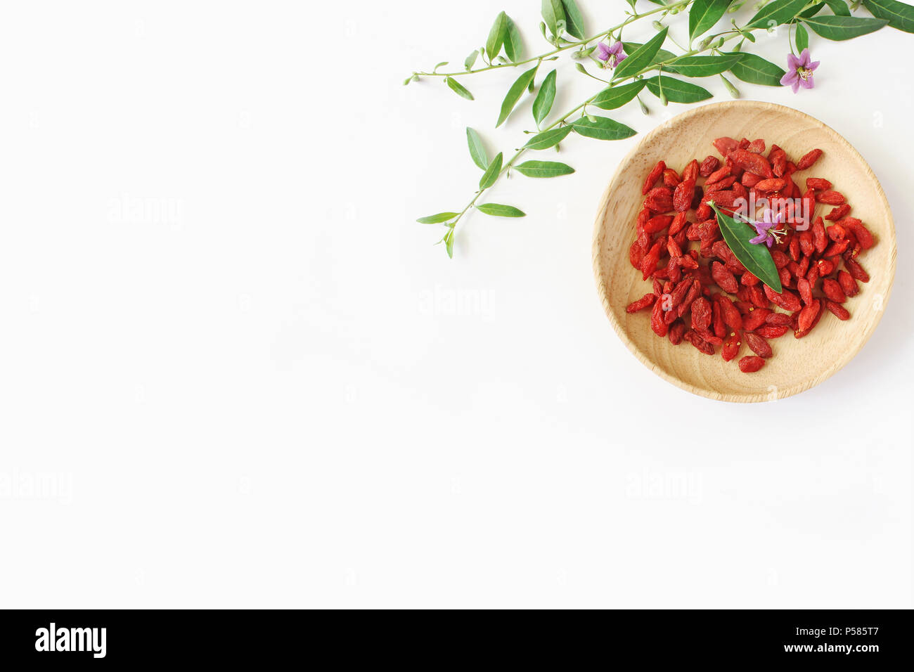 Heap of dried Goji berries fruit, Lycium barbarum in wooden bowl on white table. Blossoming wolfberry herbal plant branch. Healthy superfood. Flatlay, top view. Stock Photo