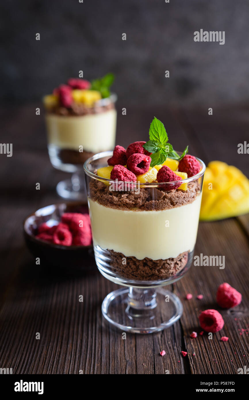 Layered mango dessert with cream cheese, crushed biscuits and freeze dried raspberries served in glass jars Stock Photo