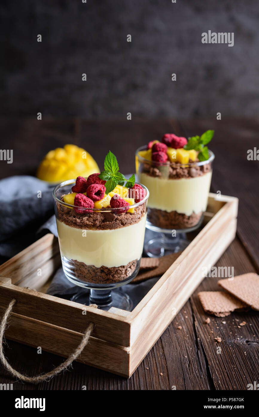 Layered mango dessert with cream cheese, crushed biscuits and freeze dried raspberries served in glass jars Stock Photo