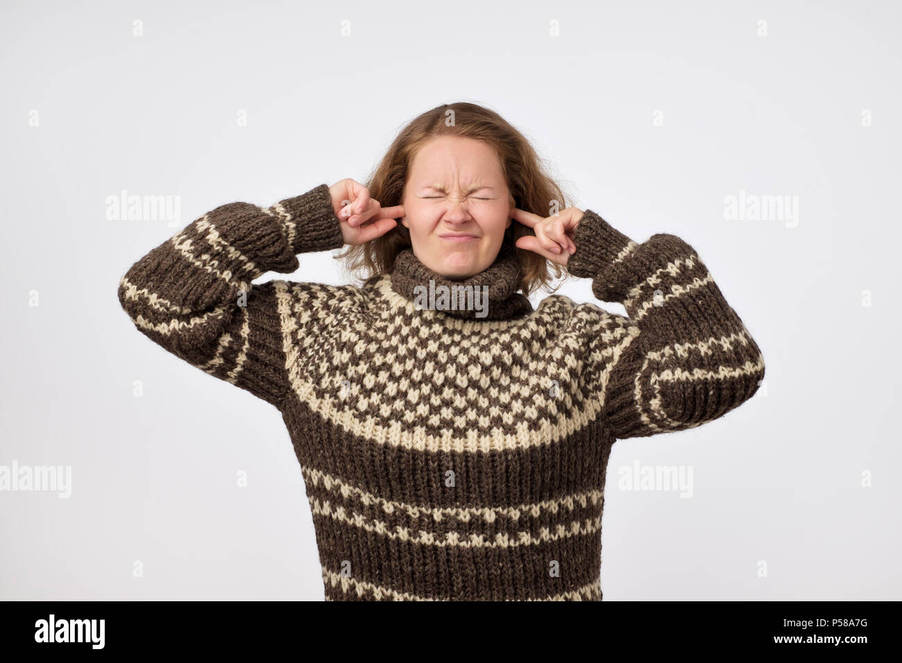 Young female in warm brown sweater plugging her ears and frowning her face being annoyed with noise. She wants to stay in calm comfortable place being Stock Photo