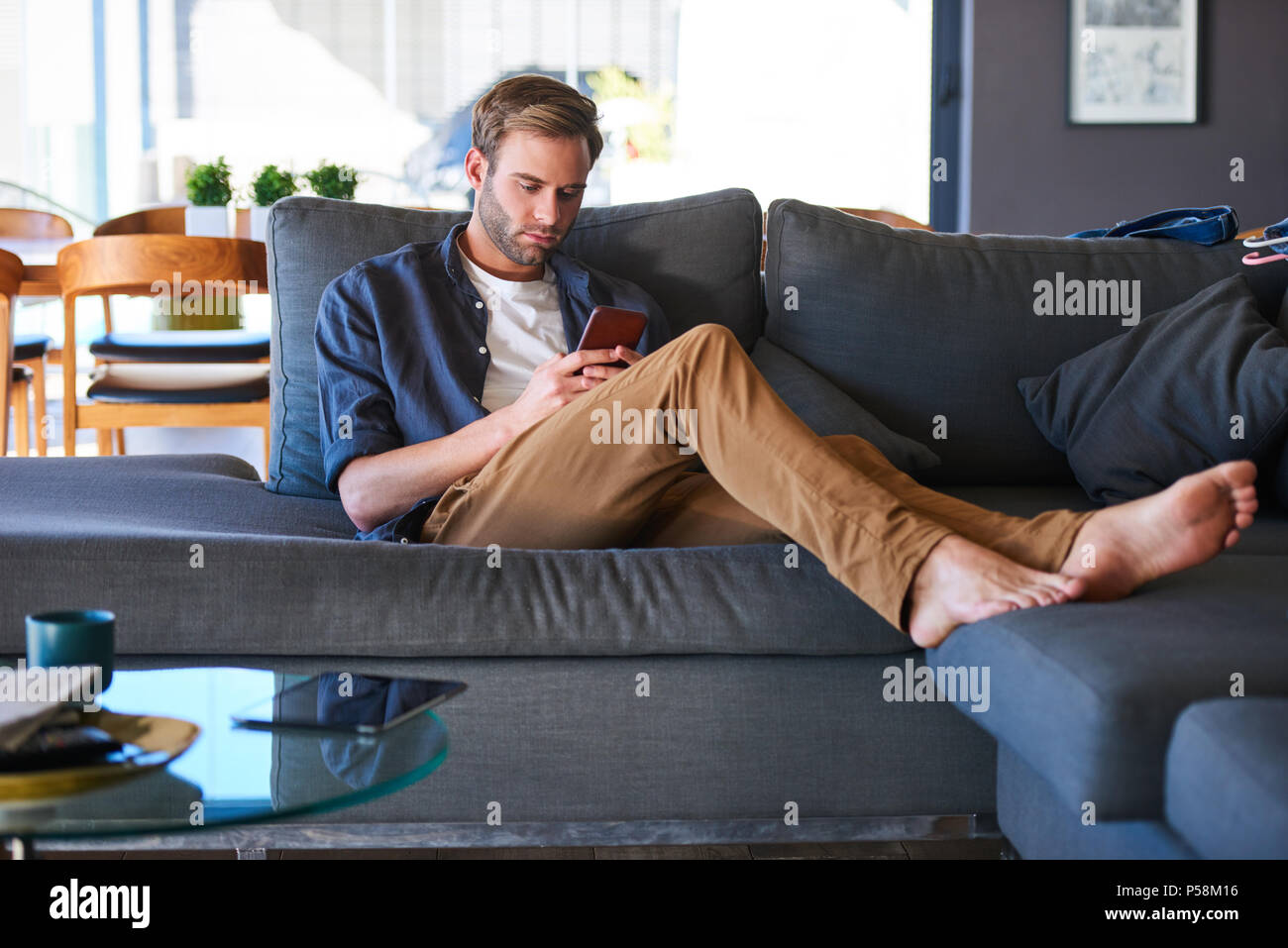 Wide shot of an attractive caucasian man sitting on a generous sofa in a modern home, while texting on his latest mobile phone with his feet up on the couch and relaxing. Stock Photo