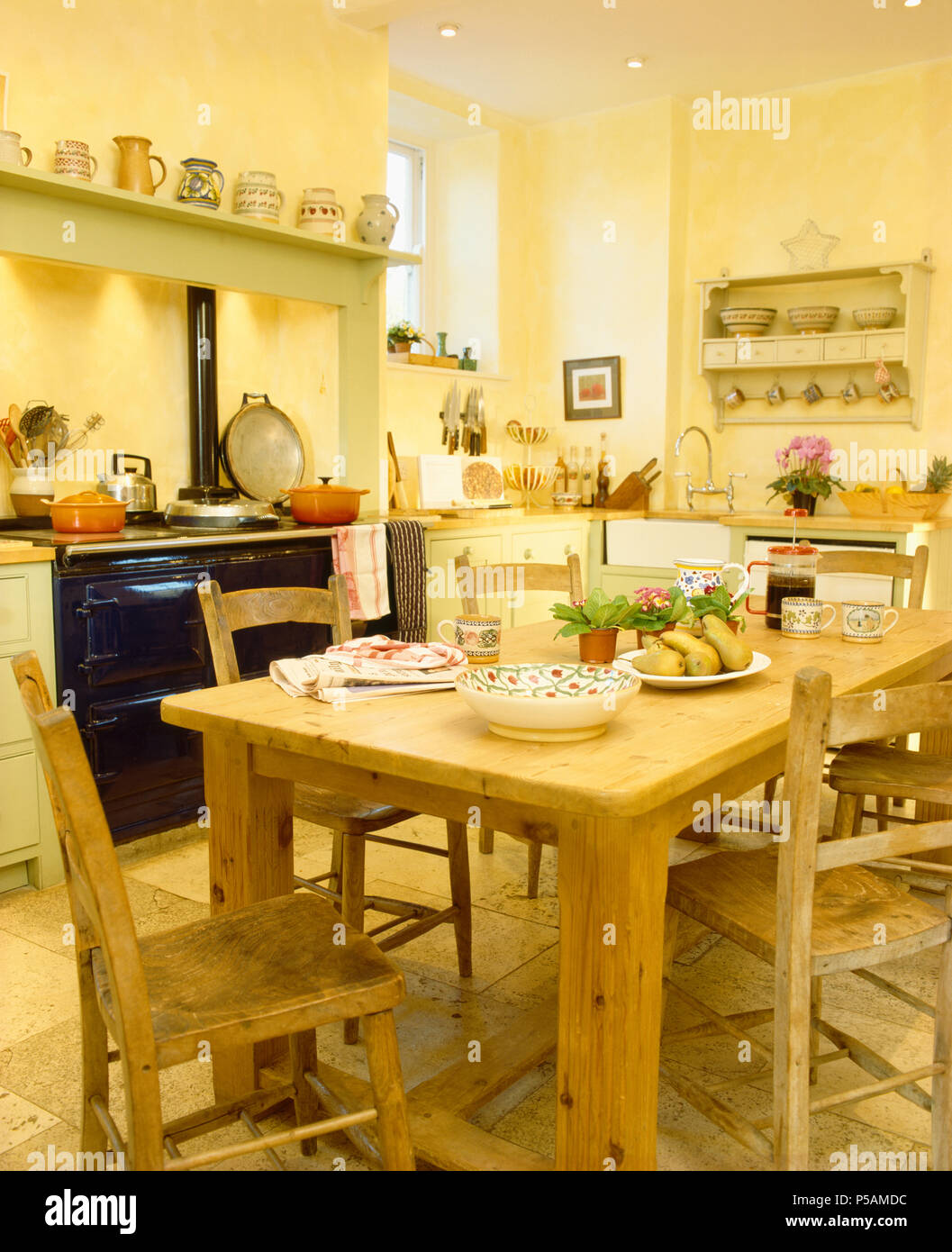 PIne chairs and heavy pine table in yellow country kitchen with black Aga Stock Photo