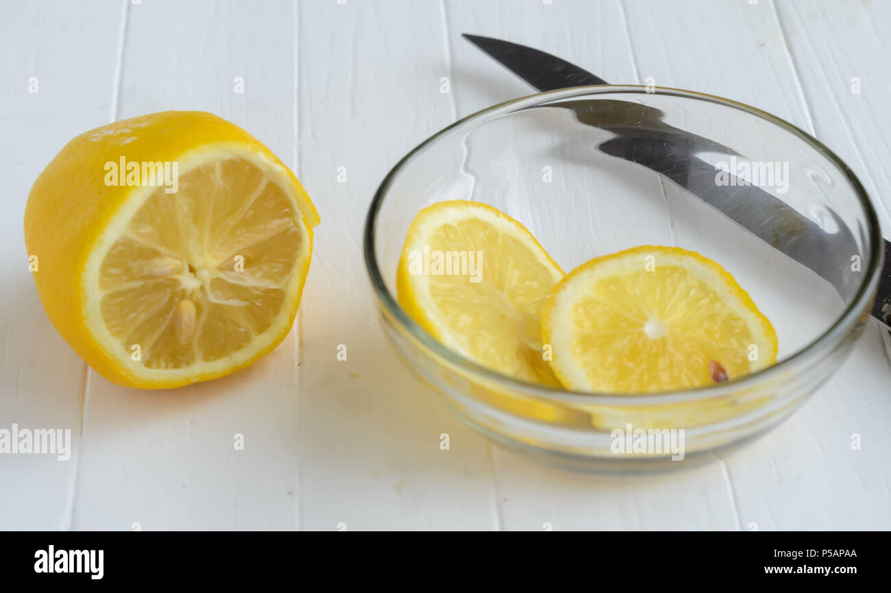 One cutted lemon and two slices of lemon into glass plate.Part of the knife blade behind the glass plate. White wooden background. Stock Photo