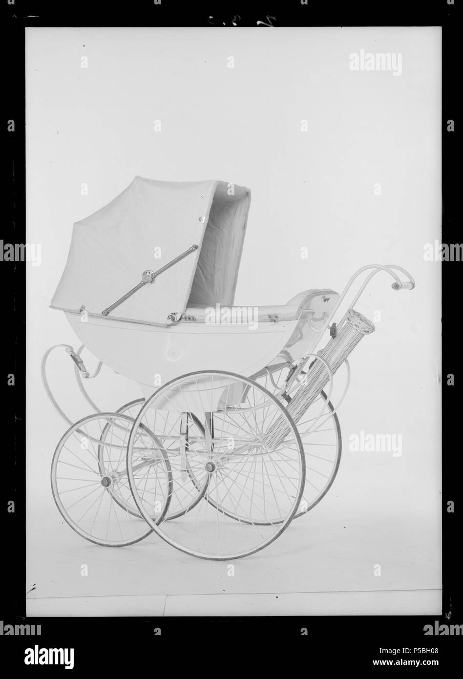 Barnvagn. Svenska: Fyrhjulig barnvagn med tvåsitsig vitlackerad kaross av  trä som vilar på en ställning av vitmålat stål. På sidorna fastbunden  undertill med vita läderremmar. Nedfällbar suflett av vaxduk med gångjärn av