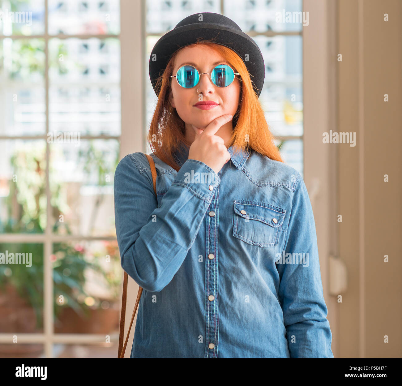Stylish redhead woman wearing bowler hat and sunglasses looking confident at the camera with smile with crossed arms and hand raised on chin. Thinking Stock Photo