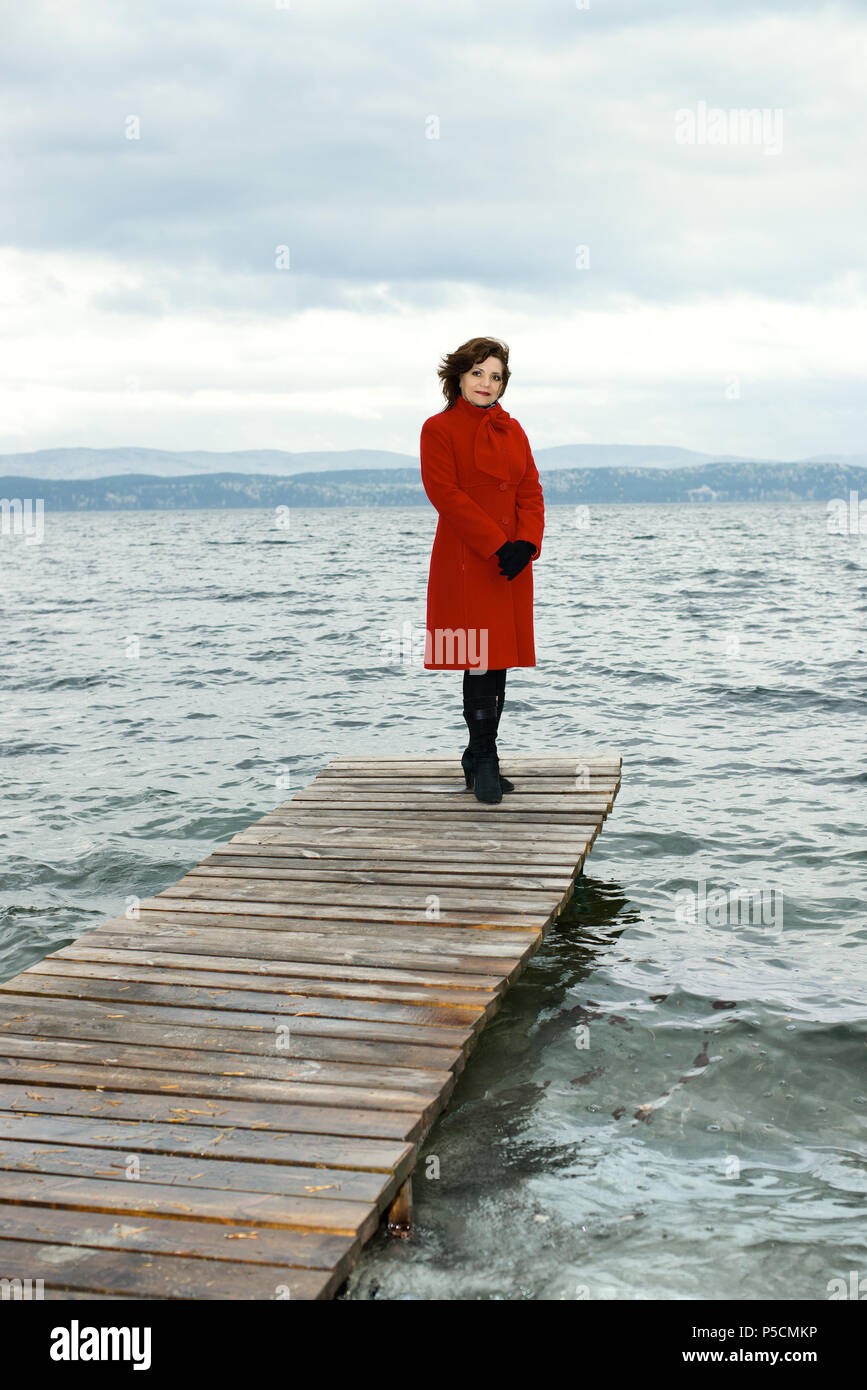 beauty mature woman in red topcoat, outdoor on water moorage for boat, in autumn cold overcast day Stock Photo