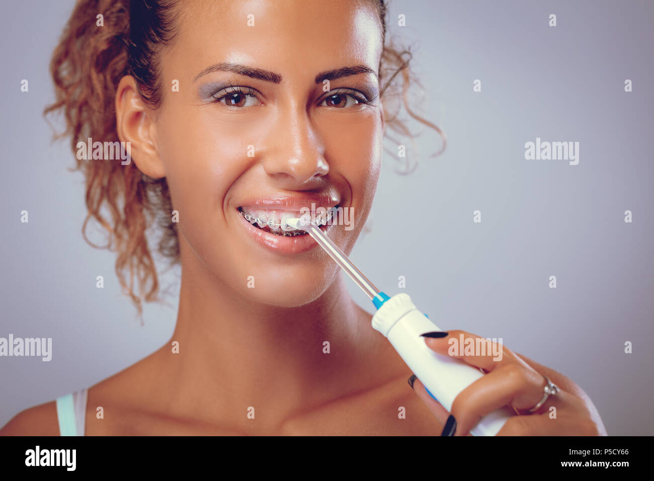 Smiling young woman with braces cleaning her teeth with water flosser. Looking at camera Stock
