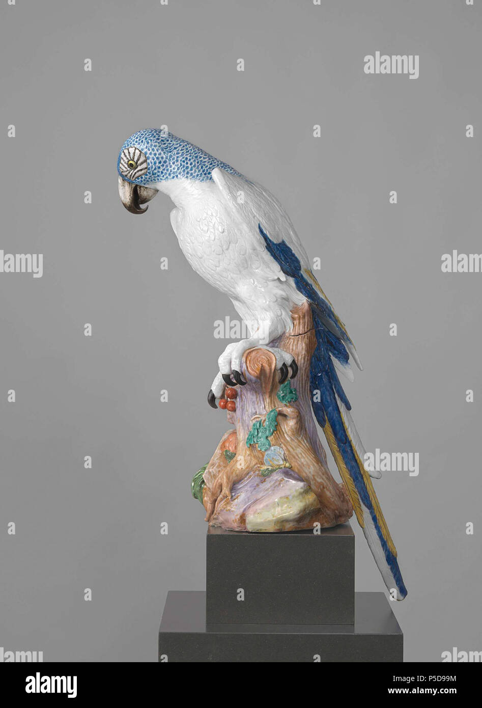 Blue Macaw . Figuur van beschilderd porselein. De stelt een papegaai (ara) voor die op een boomstronk met bladeren zit. De papegaai heeft een blauw geschubde hals en kop en een