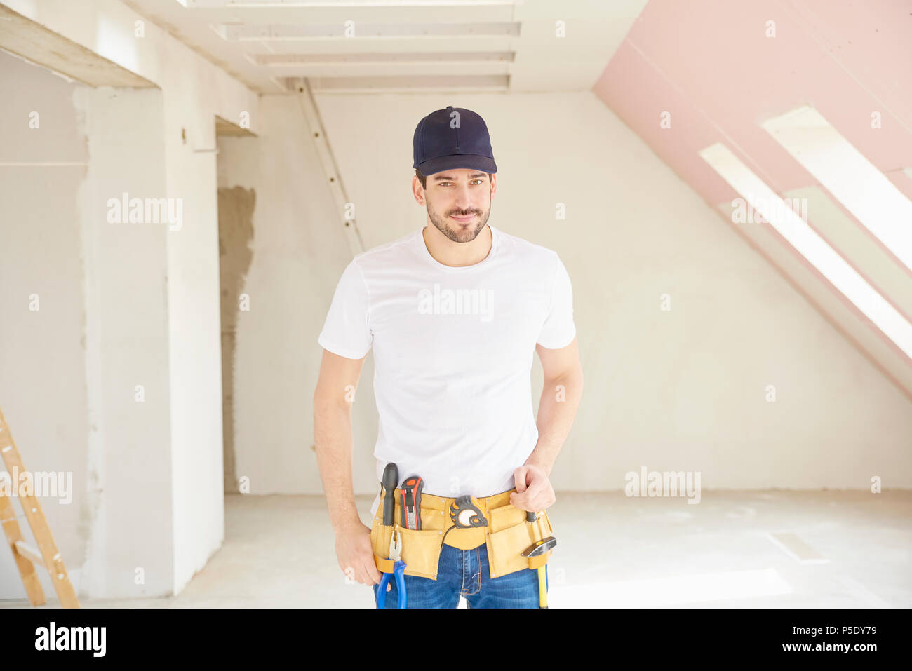 Portrait of young handyman wearing baseball cap and tool belt while standing at construction site. Stock Photo