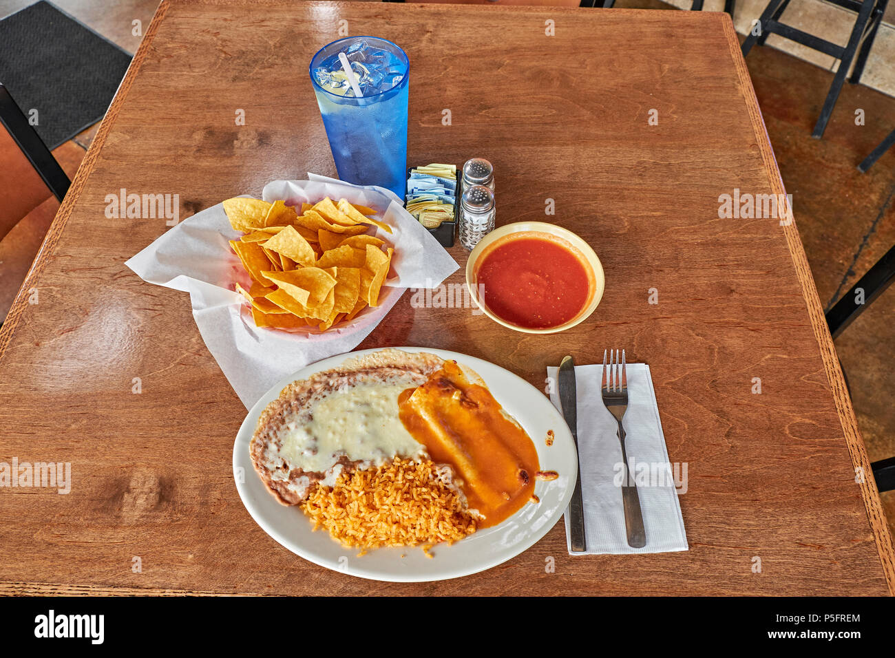 Mexican dinner plate with an enchilada, beans and rice with a basket of chips and salsa on a restaurant table top. Stock Photo
