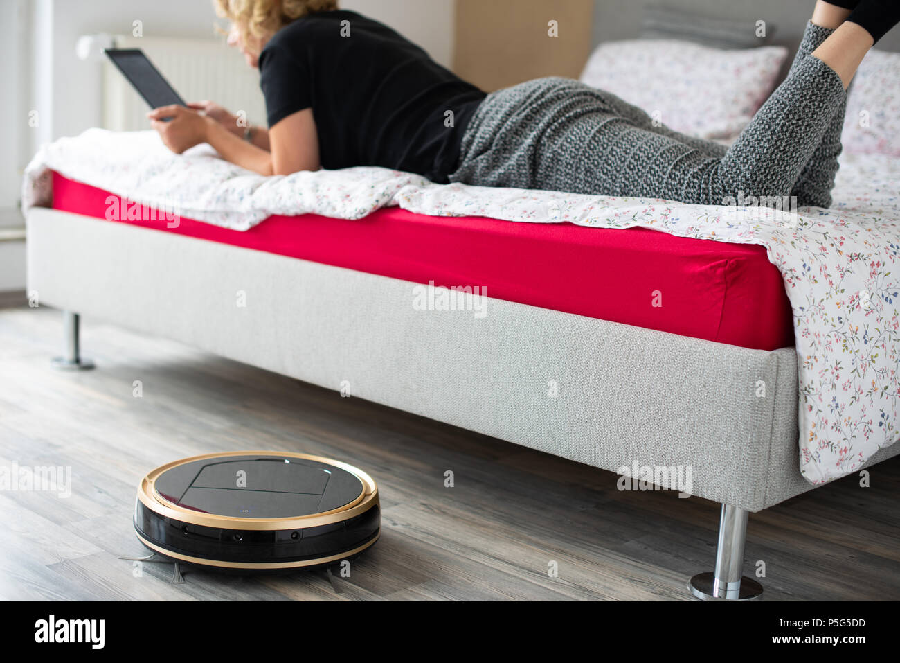 A middle aged woman is reading on her digital tablet on her bed while her vaccum cleaning robot is cleaning the wooden floor at home. Household, leisu Stock Photo